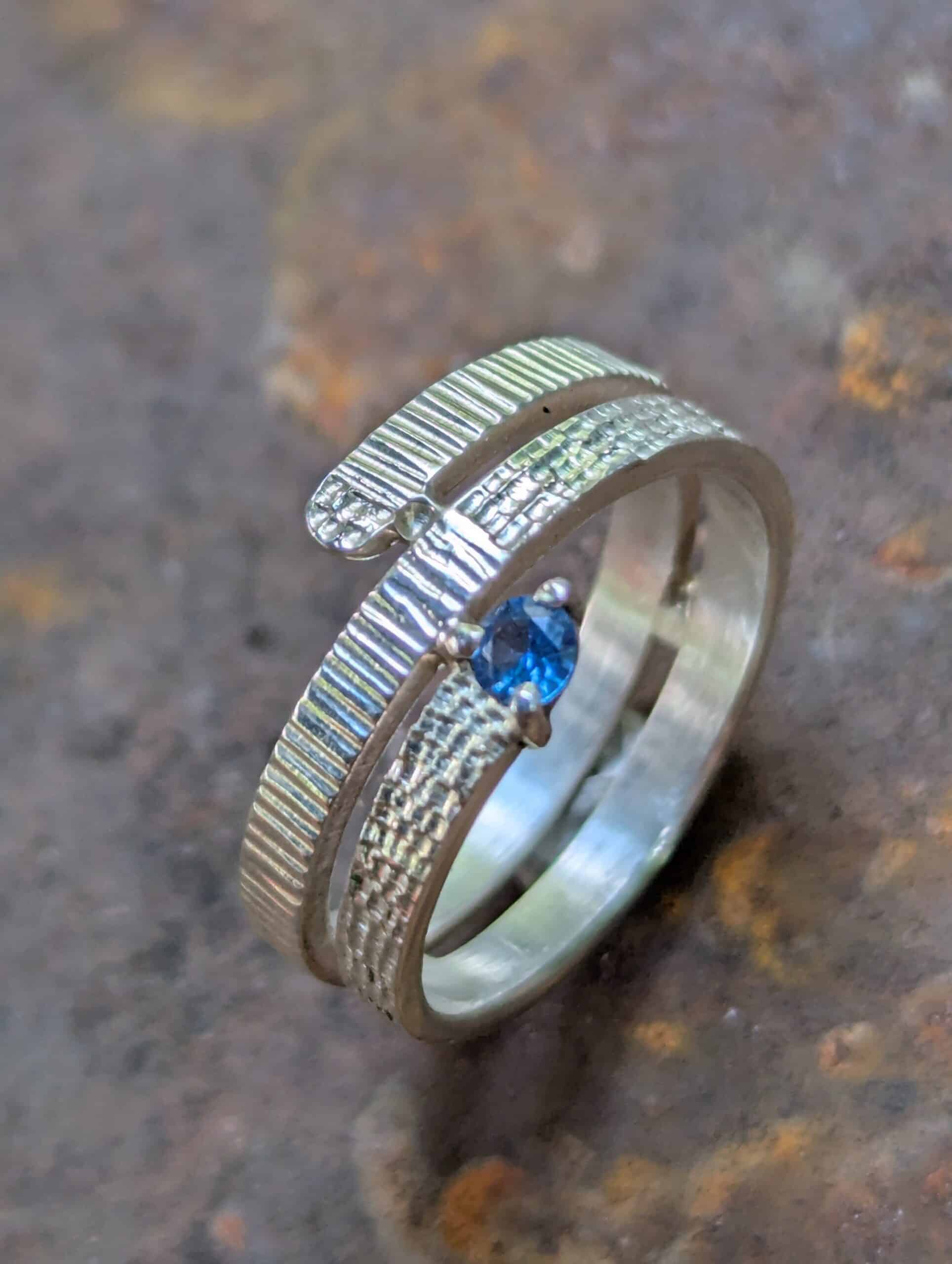 Bague Artisanale en Argent Texturé avec Pierre Bleue Bague enveloppante en argent texturé sertie d'une pierre bleue scintillante sur un fond métallique rustique.